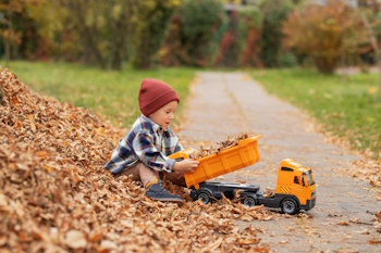 Young child sits in pile of leaves, using yellow dump truck to haul them