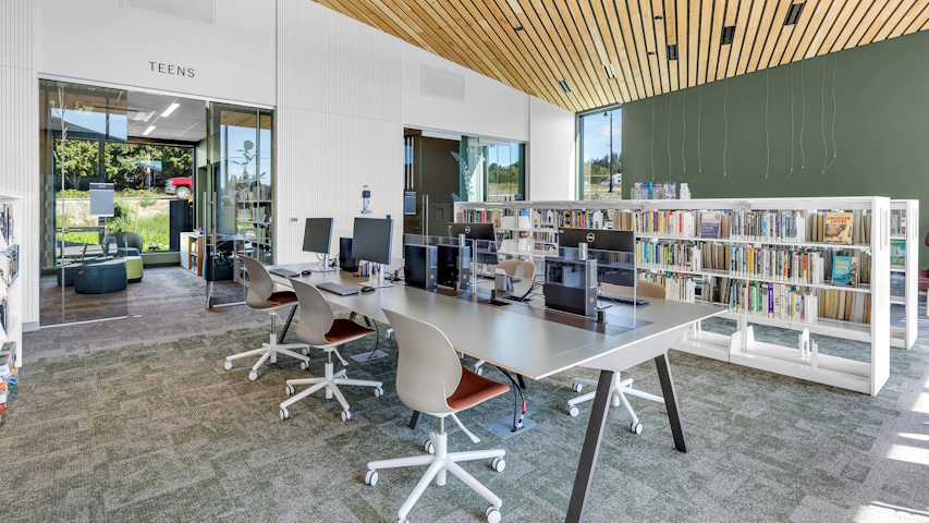 Tables with computers waiting to be used, in front of bookshelves and the doorway in to the Teen Room