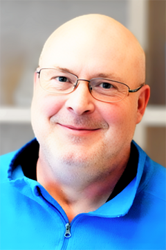 Photo of man with glasses, shaved head, wearing blue shirt, smiling